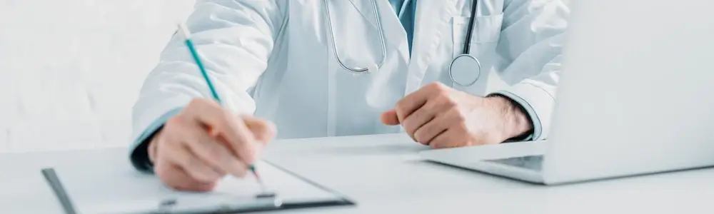 Doctor writing notes beside laptop in clinic